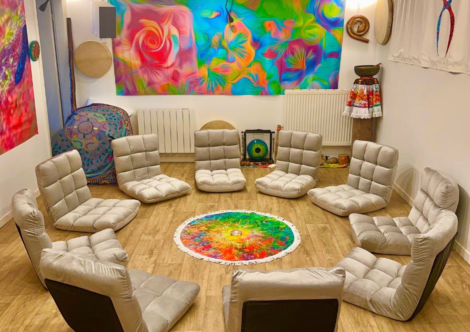 Photo d’un espace intérieur chaleureux aménagé pour les cercles d’accompagnement, avec fauteuils bas disposés en cercle autour d’un tapis mandala lumineux ; des tentures colorées et objets rituels (tambours, statuettes, offrandes, tenture Shipibo) ornent les murs — ambiance propice au sacré, à la méditation et au lien de cœur
