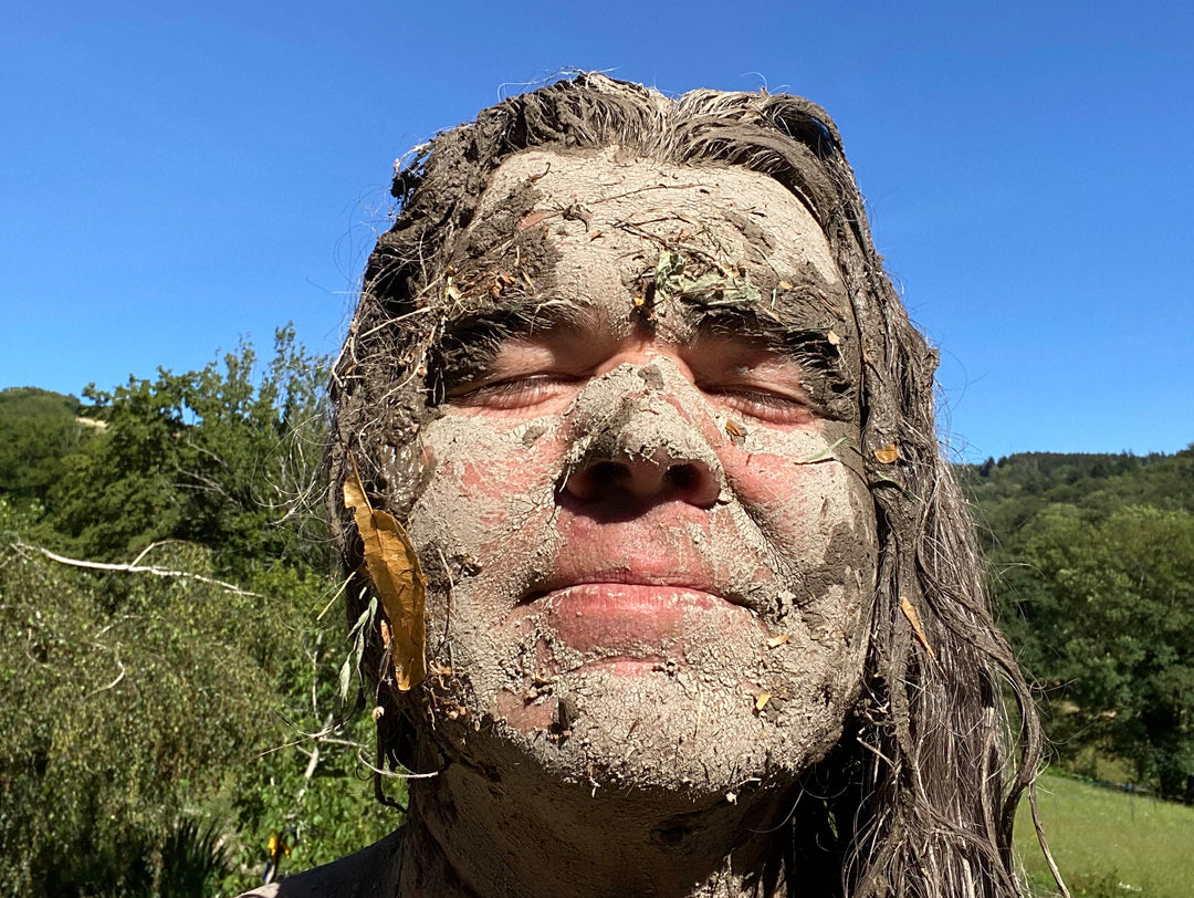 Portrait en gros plan de Xavier Gaillot, créateur d'Exstatic Designs, au visage recouvert de boue, végétaux et feuilles, les yeux fermés, baigné de lumière dans un cadre naturel verdoyant — expression de reconnexion profonde avec la Terre et les éléments