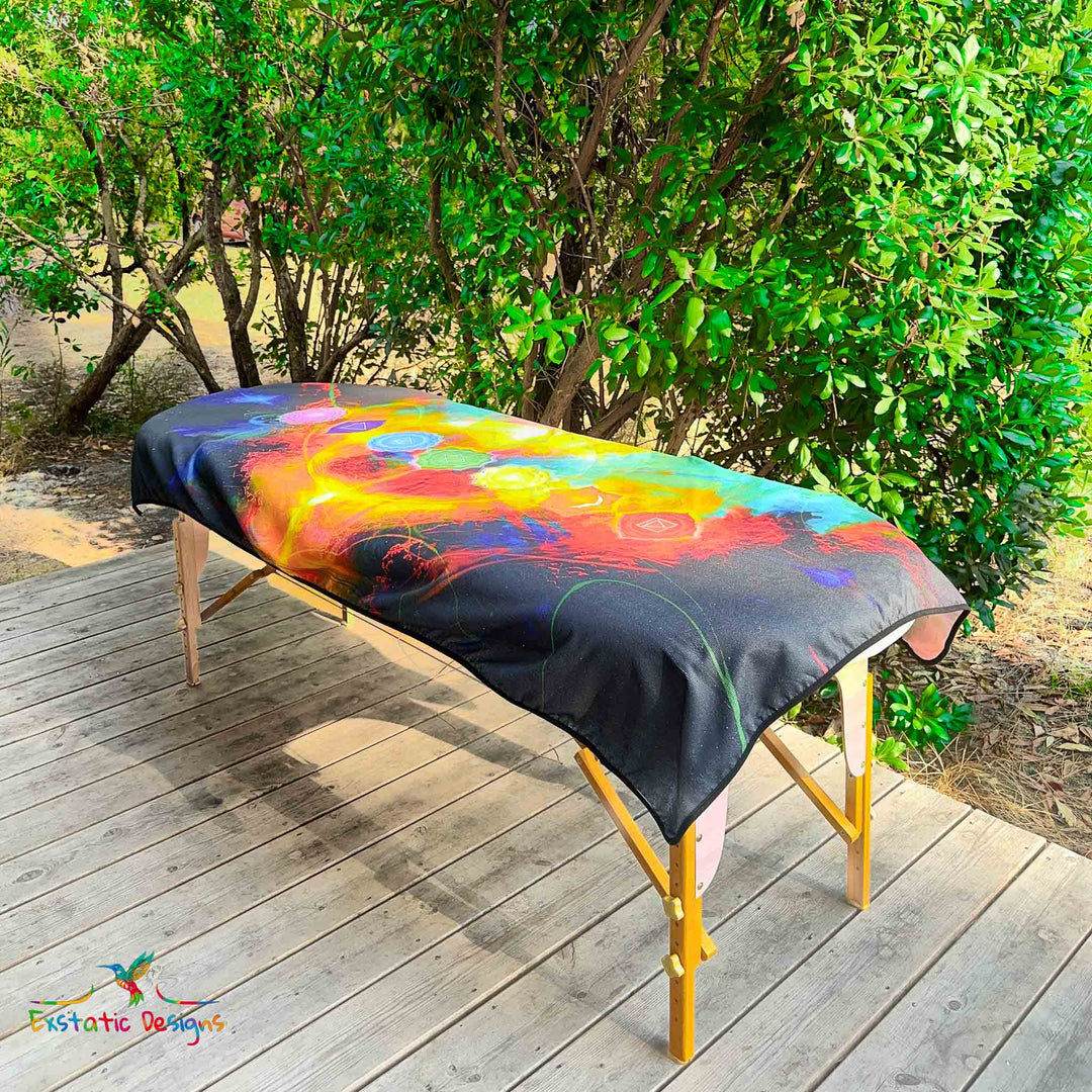 Serviette géante Tantra & Massage "L’Axe Sacré" posée sur une table de massage en extérieur, vue complète, chakras alignés verticalement sur fond arc-en-ciel cosmique, œuvre de Xavier Gaillot pour pratiques énergétiques.
