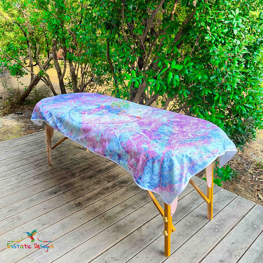 Serviette géante Tantra & Massage "Céleste Étreinte" posée sur une table de massage en extérieur, vue complète, grandes ailes angéliques et rayonnement violet-bleu, création vibratoire de Xavier Gaillot pour Exstatic Designs.