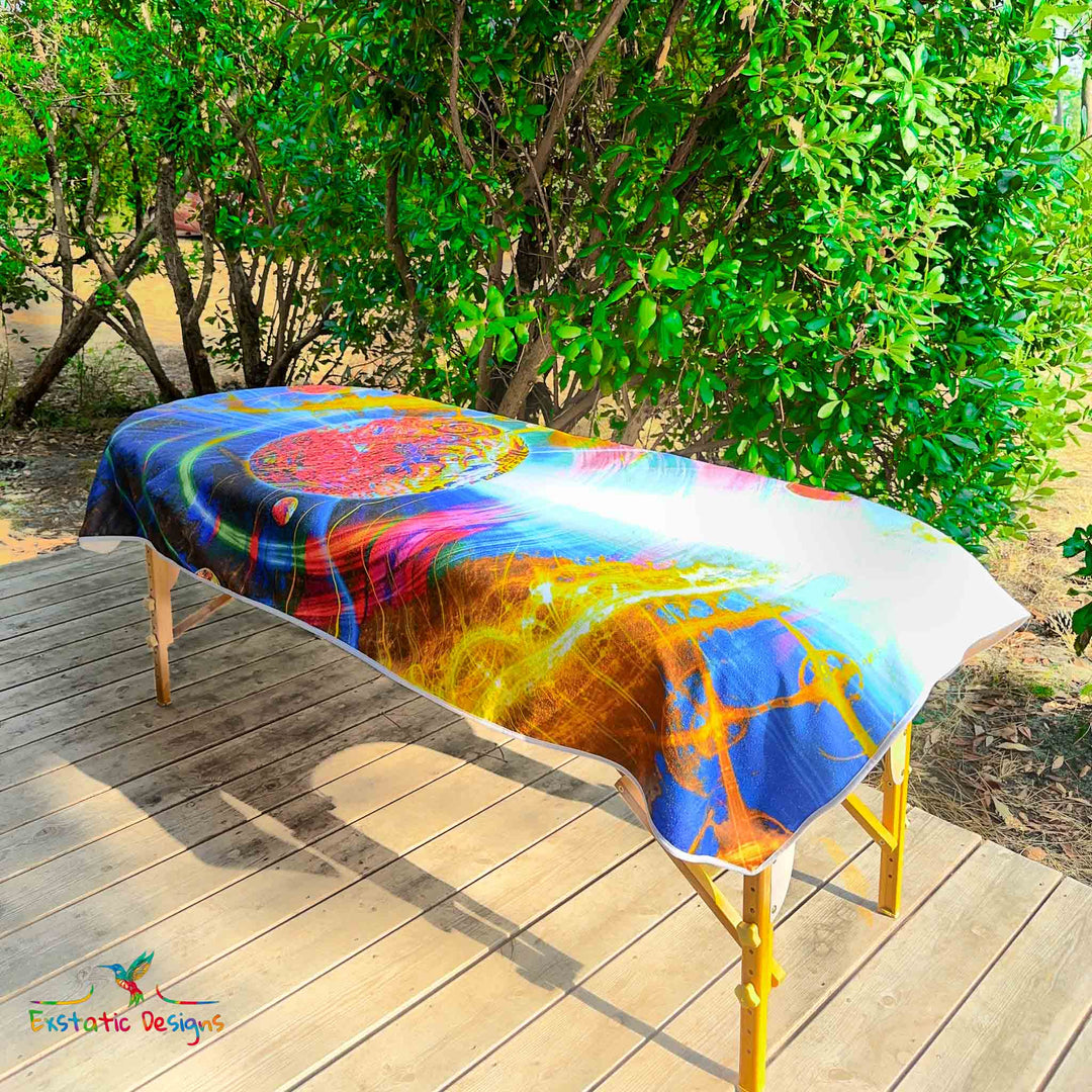 Serviette géante Tantra & Massage "La danse des sphères" posée sur une table de massage en extérieur, vue complète, design galactique avec sphères et spirales dorées, création vibratoire de Xavier Gaillot.