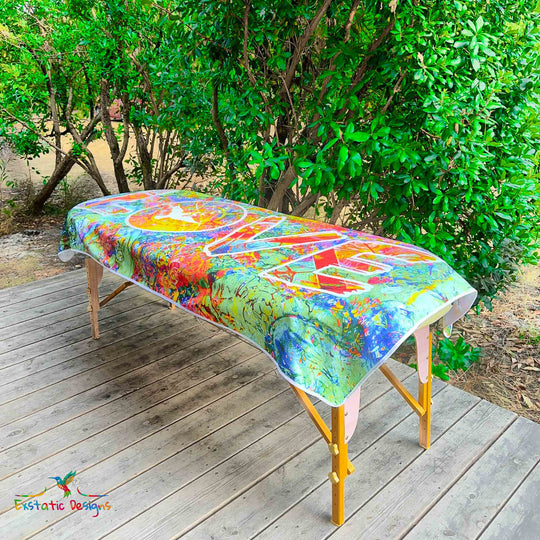 Serviette géante Tantra & Massage "Jungle d’Amour" posée sur une table de massage en extérieur, vue complète, mot LOVE vertical dans une jungle colorée avec colombe et mandala, création de Xavier Gaillot.
