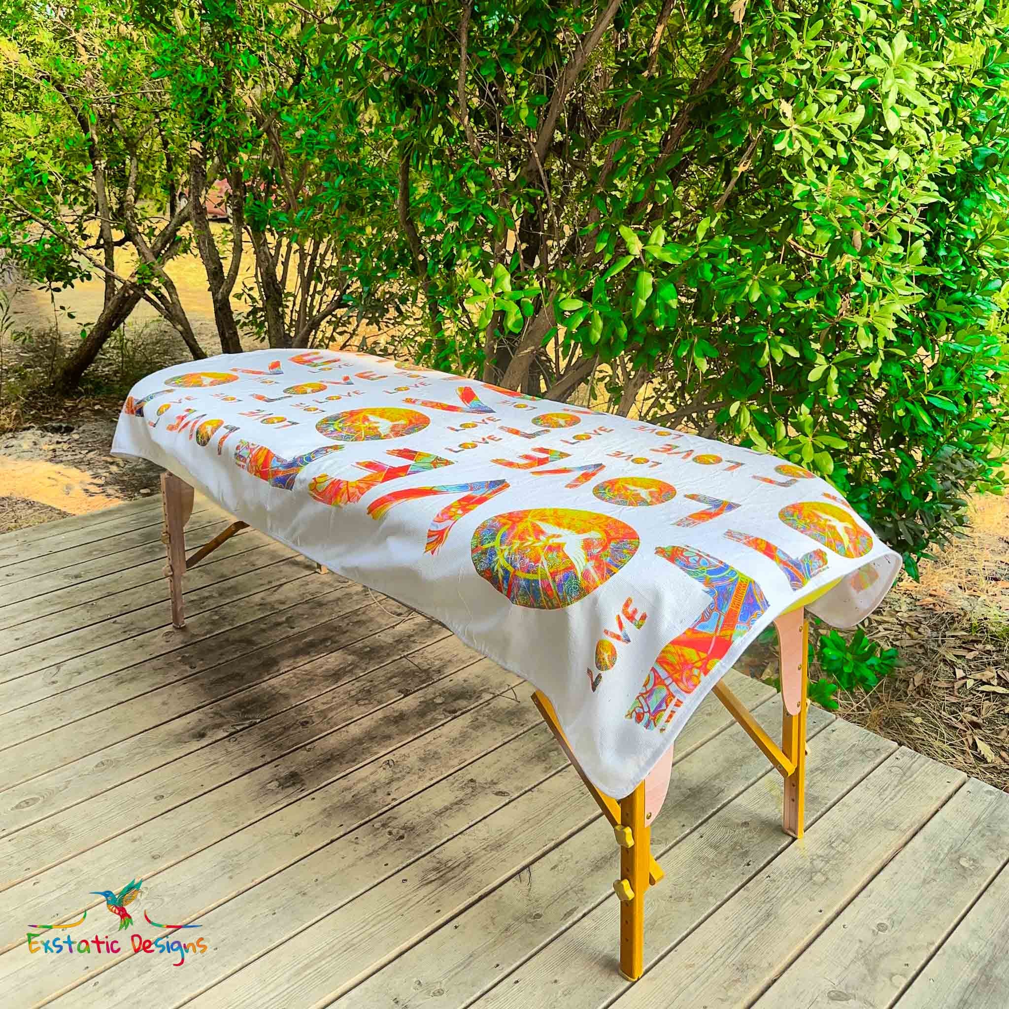 Serviette géante Tantra & Massage "L’Amour qui dégouline" posée sur une table de massage en extérieur, vue complète, motifs LOVE répétés avec colombe et mandalas solaires, création vibratoire de Xavier Gaillot.