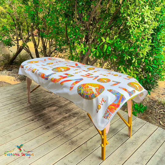 Serviette géante Tantra & Massage "L’Amour qui dégouline" posée sur une table de massage en extérieur, vue complète, motifs LOVE répétés avec colombe et mandalas solaires, création vibratoire de Xavier Gaillot.