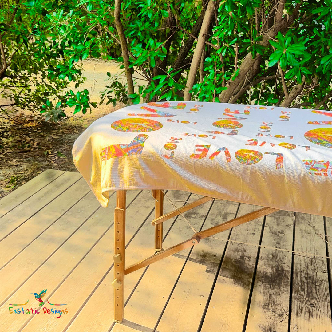 Détail de la serviette géante Tantra & Massage "L’Amour qui dégouline" sur une table de massage en extérieur, vue rapprochée des lettres LOVE et des mandalas solaires, création vibratoire de Xavier Gaillot.