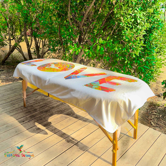 Serviette géante Tantra & Massage "L’Amour à Grande Échelle" posée sur une table de massage en extérieur, vue complète, LOVE vertical avec colombe et mandala lumineux, création vibratoire de Xavier Gaillot.