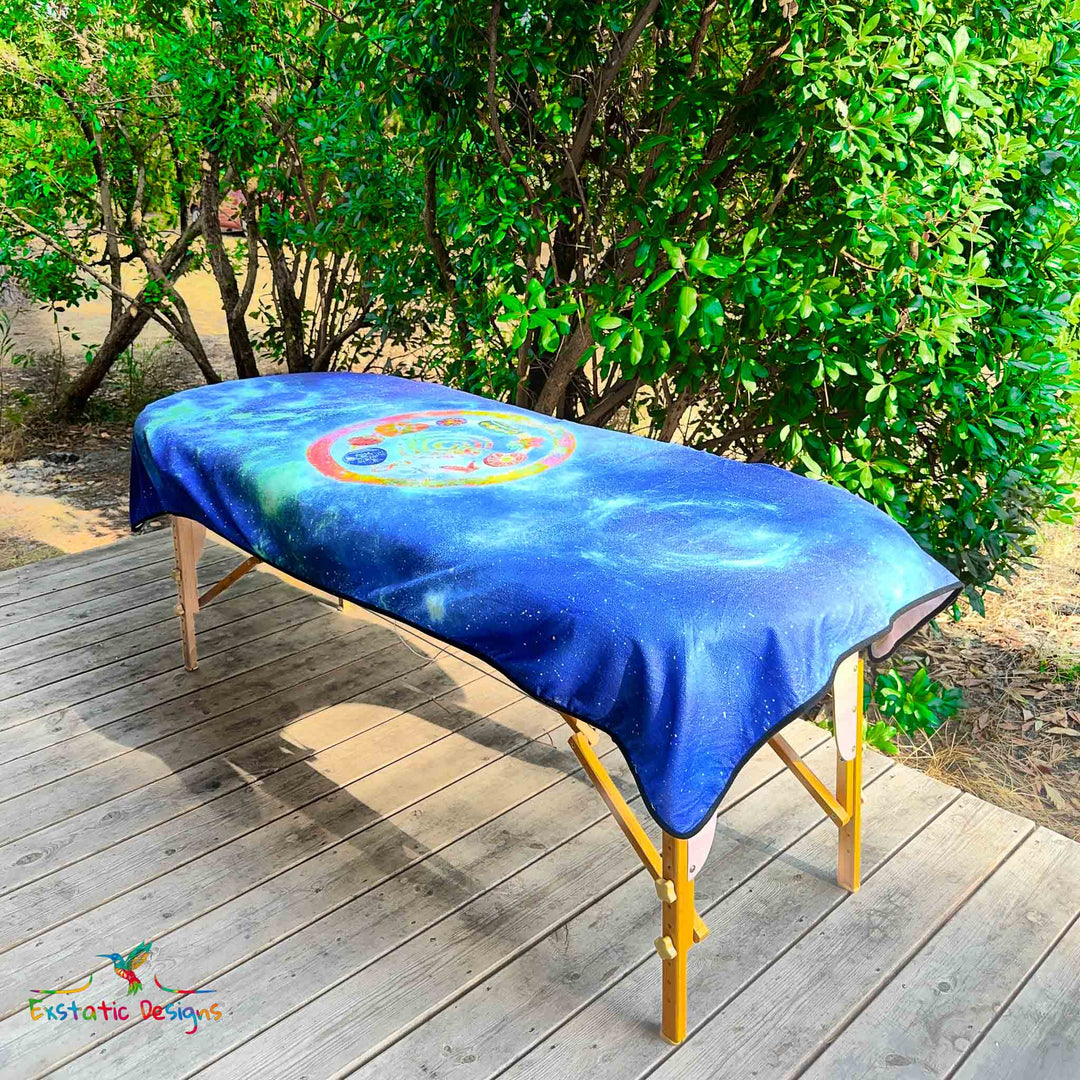 Serviette géante Tantra & Massage "Céleste mémoire" posée sur une table de massage en extérieur, vue complète, mandala galactique avec spirale, étoiles et pierres rituelles, création vibratoire de Xavier Gaillot.