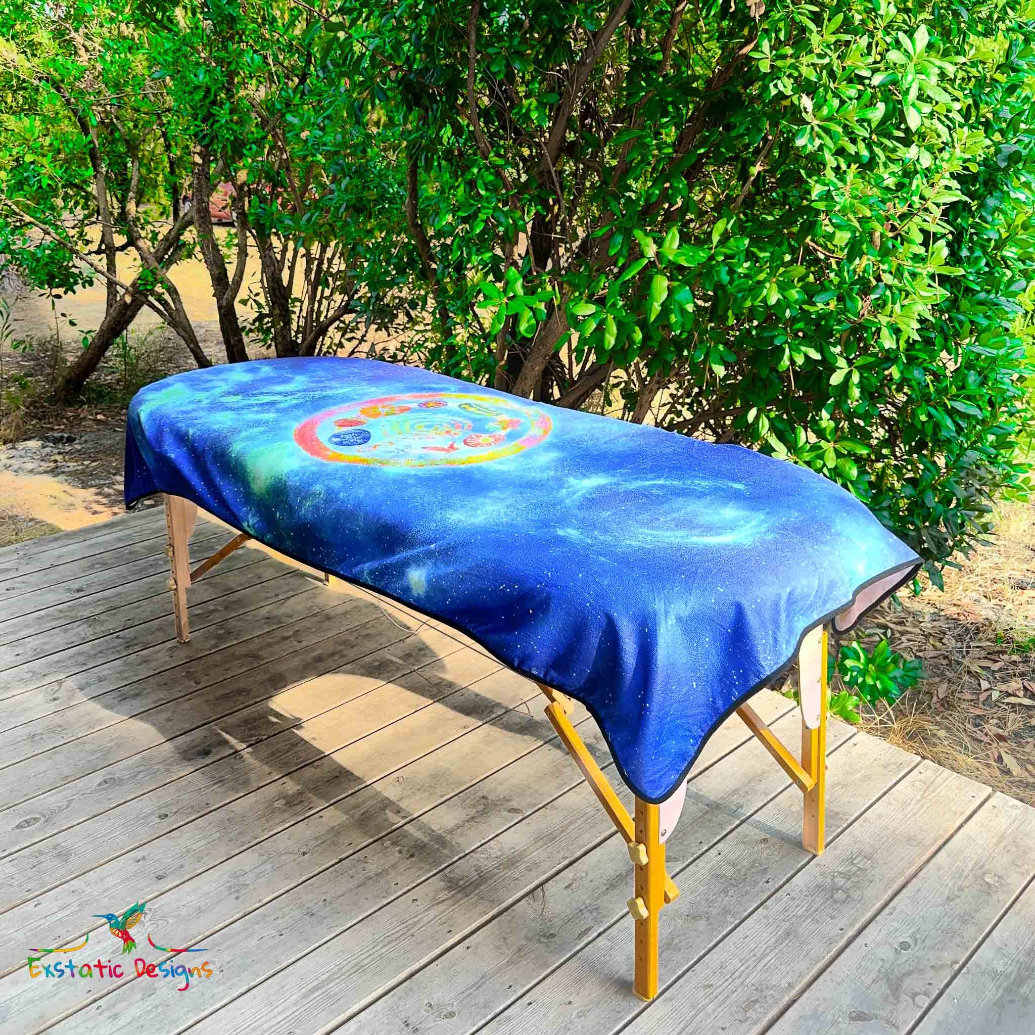 Serviette géante Tantra & Massage "Céleste mémoire" posée sur une table de massage en extérieur, vue complète, mandala galactique avec spirale, étoiles et pierres rituelles, création vibratoire de Xavier Gaillot.