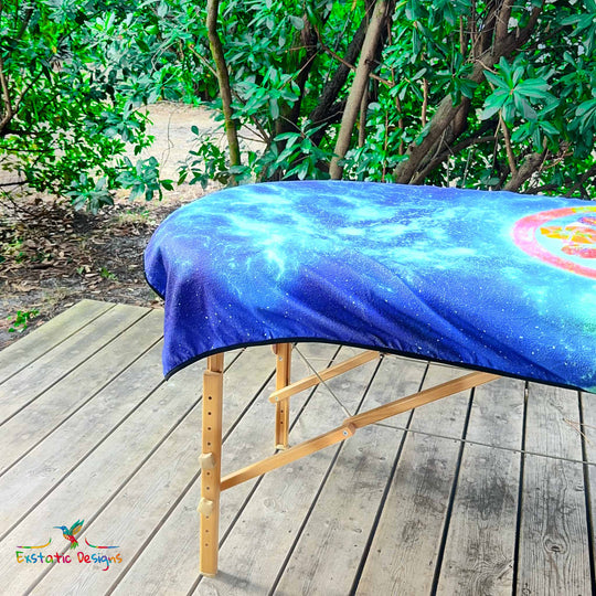 Détail de la serviette géante Tantra & Massage "Céleste mémoire" sur une table de massage en extérieur, vue rapprochée du mandala galactique et des pierres rituelles peintes, création vibratoire de Xavier Gaillot.