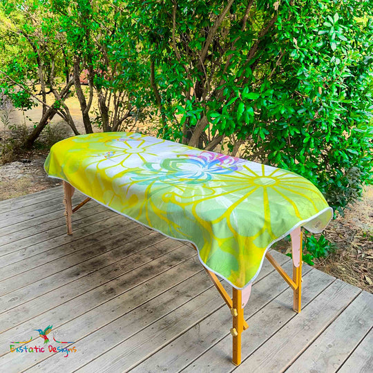 Serviette géante Tantra & Massage "Éclat de Grâce" posée sur une table de massage en extérieur, vue complète, colibris multicolores, lotus rayonnant et motifs floraux lumineux sur fond jaune, création spirituelle de Xavier Gaillot.