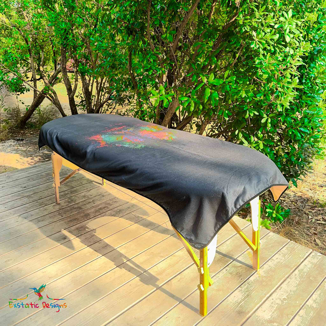 Serviette géante Tantra & Massage "La magie du papillon" posée sur une table de massage en extérieur, vue complète, papillon mystique et coloré surgissant d’un fond profond, œuvre vibratoire de Xavier Gaillot.