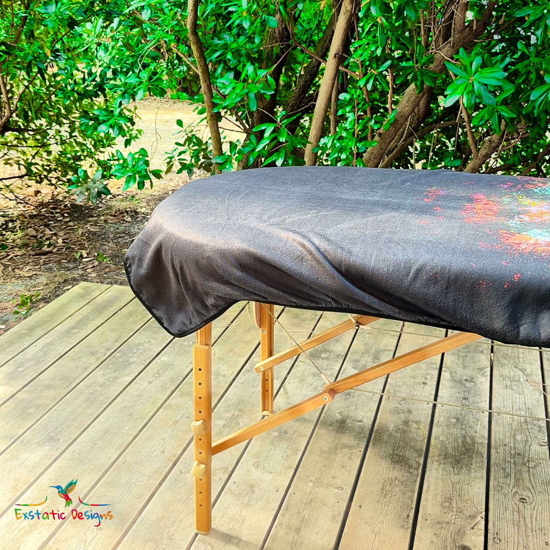 Détail de la serviette géante Tantra & Massage "La magie du papillon" sur une table de massage en extérieur, vue rapprochée du papillon coloré et des volutes lumineuses, création vibratoire de Xavier Gaillot.