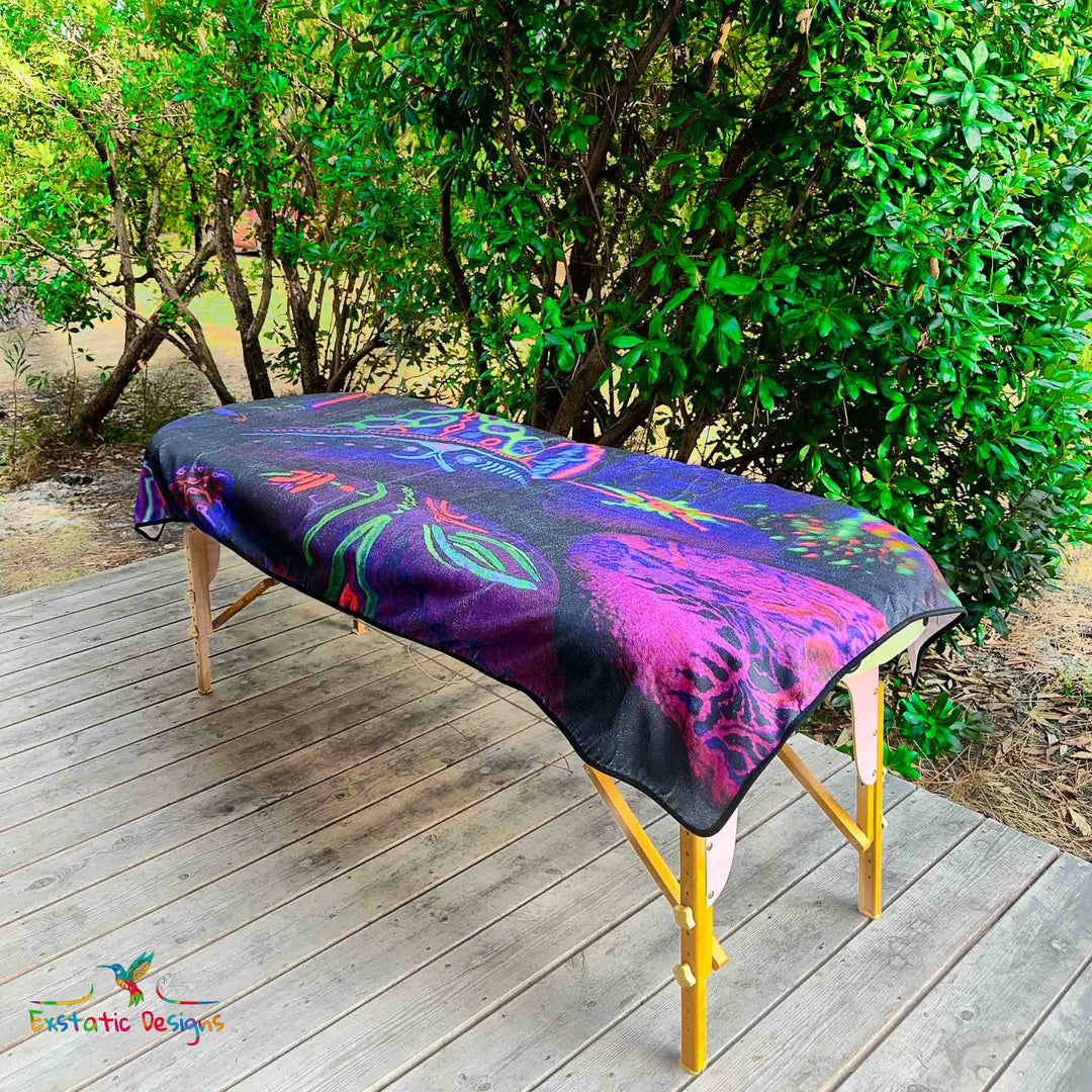 Serviette géante Tantra & Massage "Mémoires initiatiques" posée sur une table de massage en extérieur, vue complète, pierres rituelles peintes à la main et lumière noire UV évoquant la mémoire minérale, création sacrée de Xavier Gaillot.