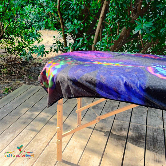 Détail de la serviette géante Tantra & Massage "Mémoires initiatiques" sur une table de massage en extérieur, vue rapprochée des pierres rituelles peintes et des reflets lumineux, création sacrée de Xavier Gaillot.