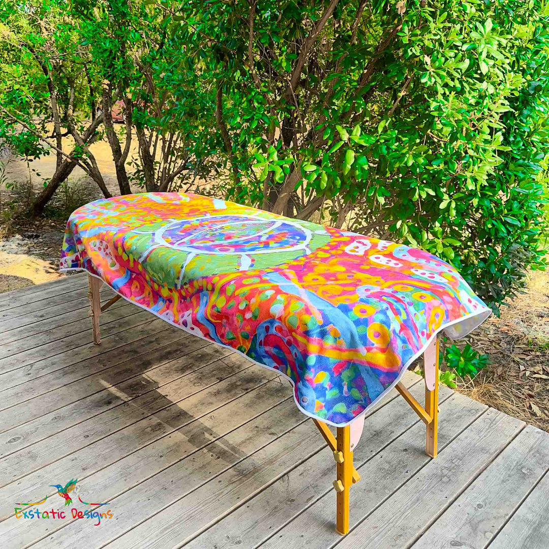 Serviette géante Tantra & Massage "Ouvre l’Œil" posée sur une table de massage en extérieur, vue complète, œil magique central et couleurs vives, création vibratoire de Xavier Gaillot pour Exstatic Designs.