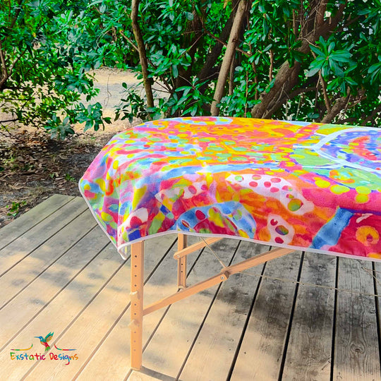 Détail de la serviette géante Tantra & Massage "Ouvre l’Œil" sur une table de massage en extérieur, vue rapprochée de l’œil central et des motifs colorés, création vibratoire de Xavier Gaillot pour Exstatic Designs.