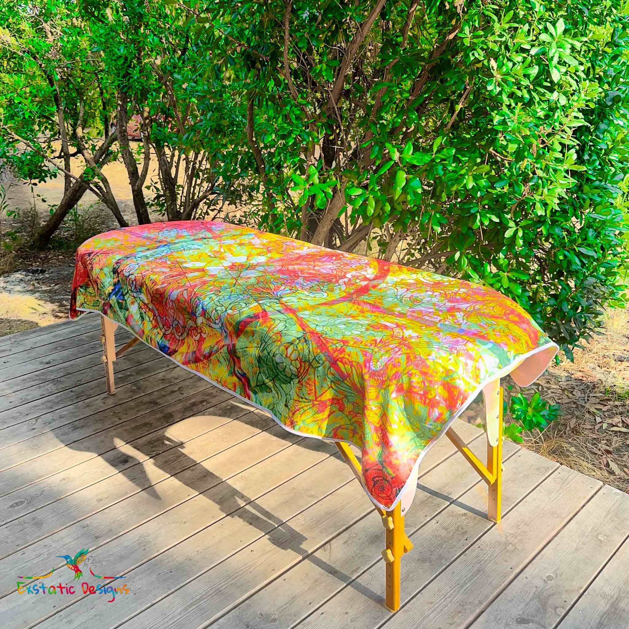 Serviette géante Tantra & Massage "Sacrée grâce" posée sur une table de massage en extérieur, vue complète, mandala floral style vitrail sacré baigné de lumière, création lumineuse de Xavier Gaillot.