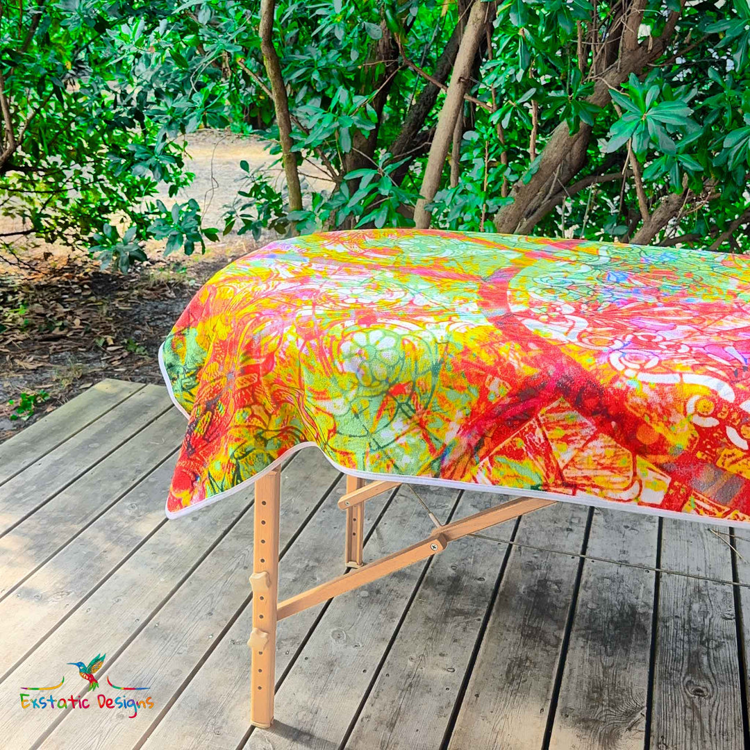 Détail de la serviette géante Tantra & Massage "Sacrée grâce" sur une table de massage en extérieur, vue rapprochée du mandala floral et des effets de vitrail sacré, création lumineuse de Xavier Gaillot.
