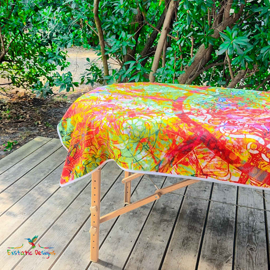 Détail de la serviette géante Tantra & Massage "Sacrée grâce" sur une table de massage en extérieur, vue rapprochée du mandala floral et des effets de vitrail sacré, création lumineuse de Xavier Gaillot.