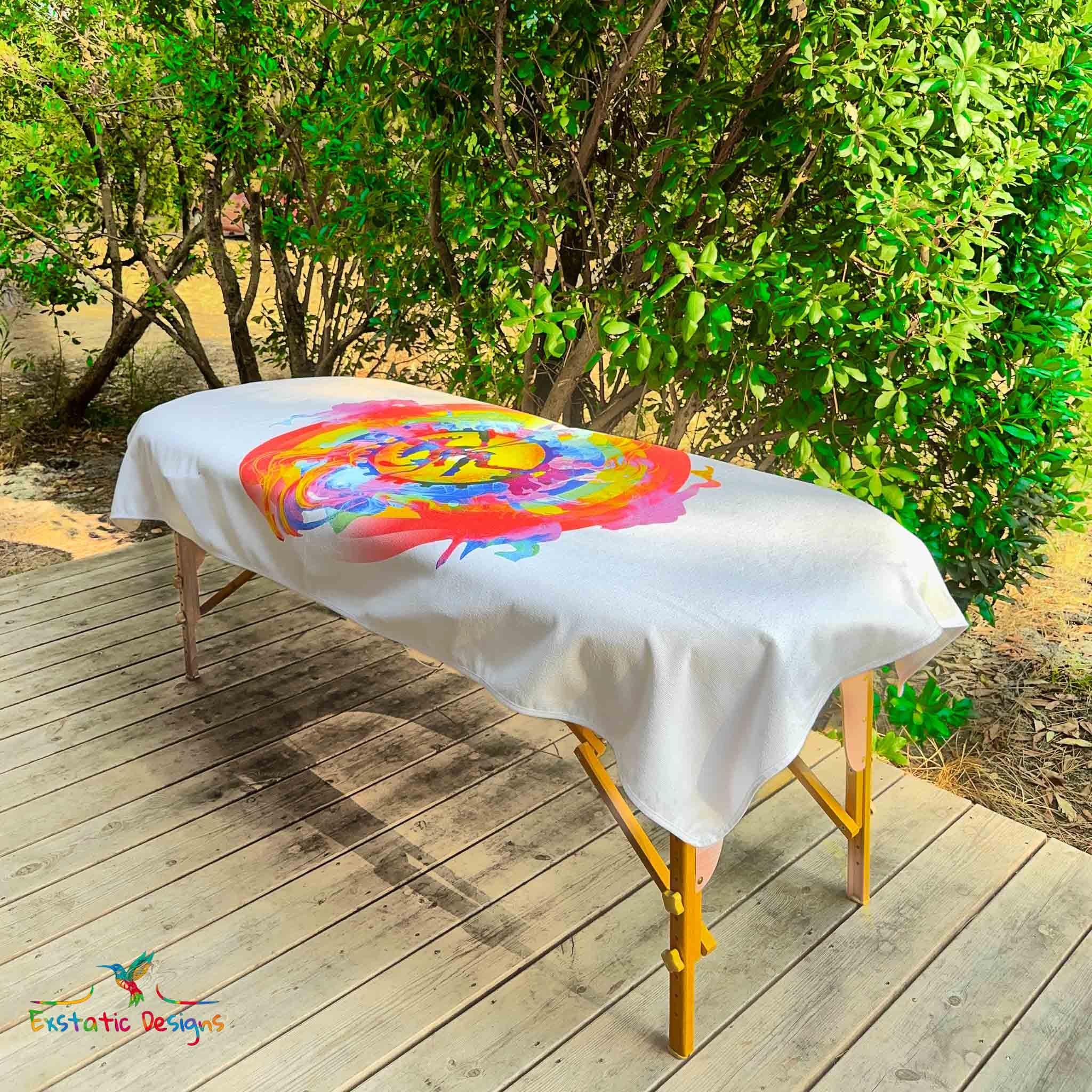 Serviette géante Tantra & Massage "Shiva danse" posée sur une table de massage en extérieur, vue complète, symbole sacré de Shiva entouré d’un halo arc-en-ciel et de volutes colorées, création spirituelle de Xavier Gaillot.