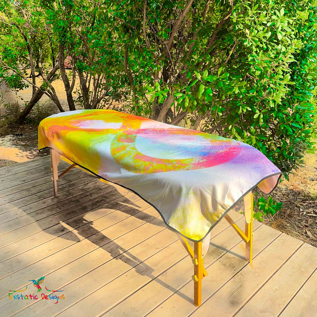 Serviette géante Tantra & Massage "Le Souffle de l’Infini" posée sur une table de massage en extérieur, vue complète, symbole de l’infini au centre d’un halo coloré vibrant, création spirituelle de Xavier Gaillot.